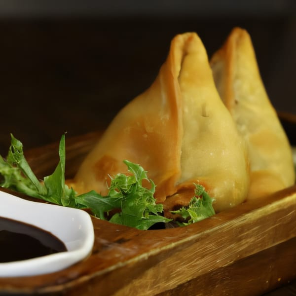 Two golden-brown Nepalese samosas with green lettuce on a wooden tray, with a side of dark sauce.