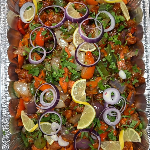 Nepalese chili chicken with bell peppers, onions, cilantro, and lemon slices in a foil tray.